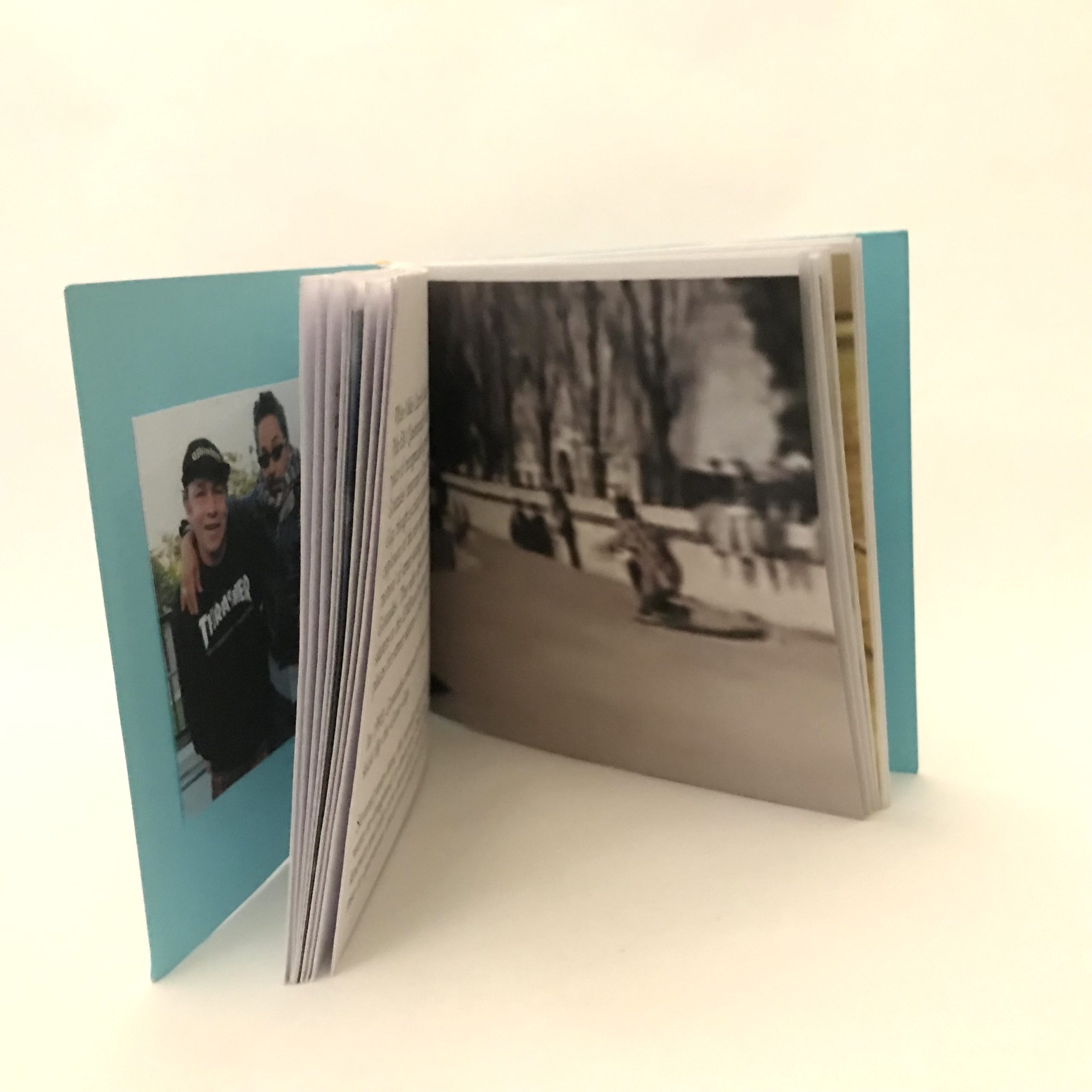 Artist's book standing upright and open on white background. On the front inside cover is a photograph of two people. The person on the left is wearing a black baseball hat and sweater with the words 'Thrasher' in white. The person on the right has their arm around the person on the left. They are wearing sunglasses, a scarf and a leather jacket. Other opened pages depict unreadable text and a screen capture of a person compressed on a skateboard after landing a trick. People in the background are standing and sitting on ledges observing the trick.