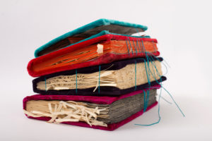 Four books of multiple colors stacked on top of each other, the covers are made of velvet and the pages are frayed. There are teal colored threads coming out of the second book, which is red with orange pages