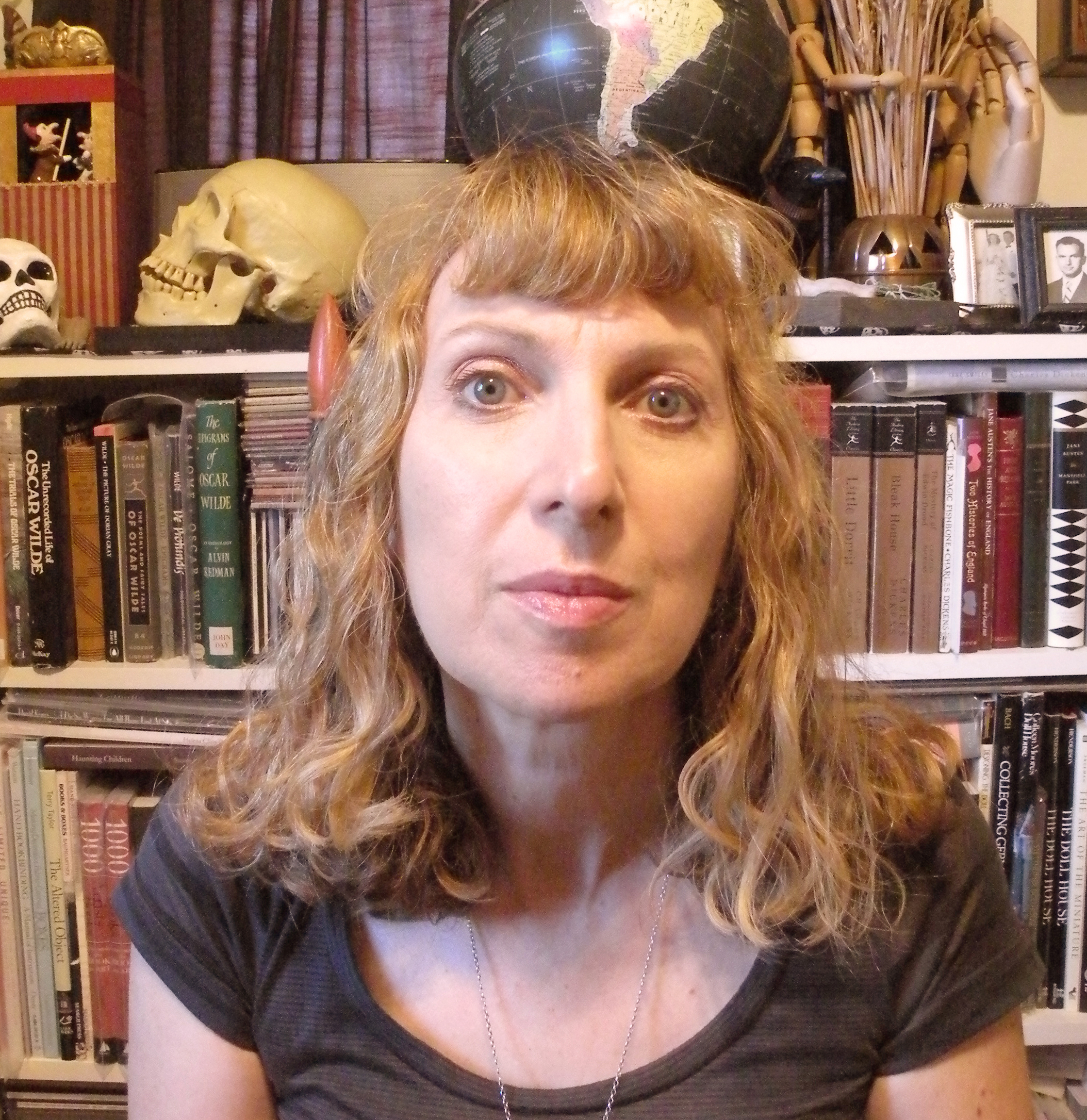 Eileen, a white woman with curly strawberry-blone hair and bangs, looks at the camera in front of a bookcase full of books and objects including a skull and a globe.