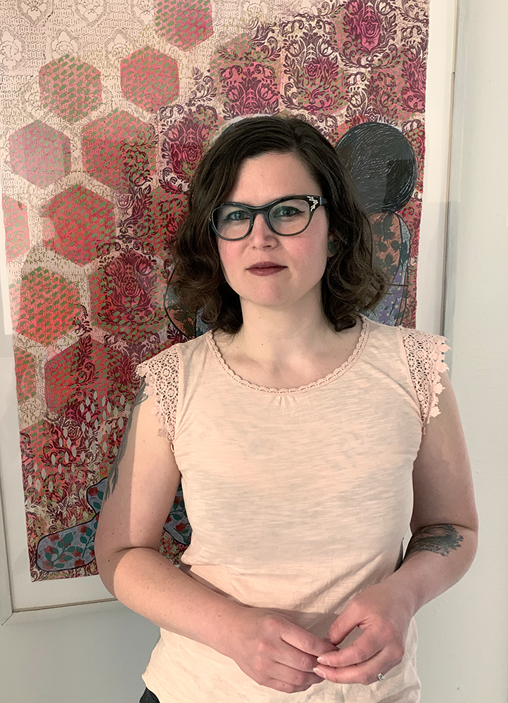 Liz stands in front of her artwork. She is a white woman with glasses, should length wavy brown hair wearing a light pink shirt.