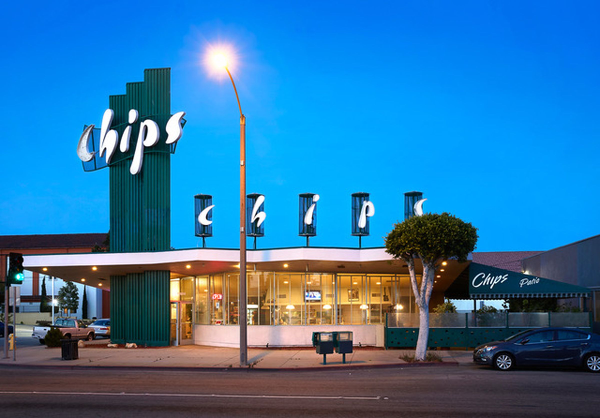 Image of Chips Restaurant: Mid century diner with the word "CHIPS" across the top. The sky surrounding the diner is dim, there is a bright street light in front of the building. The lights inside of the building are all turned on.