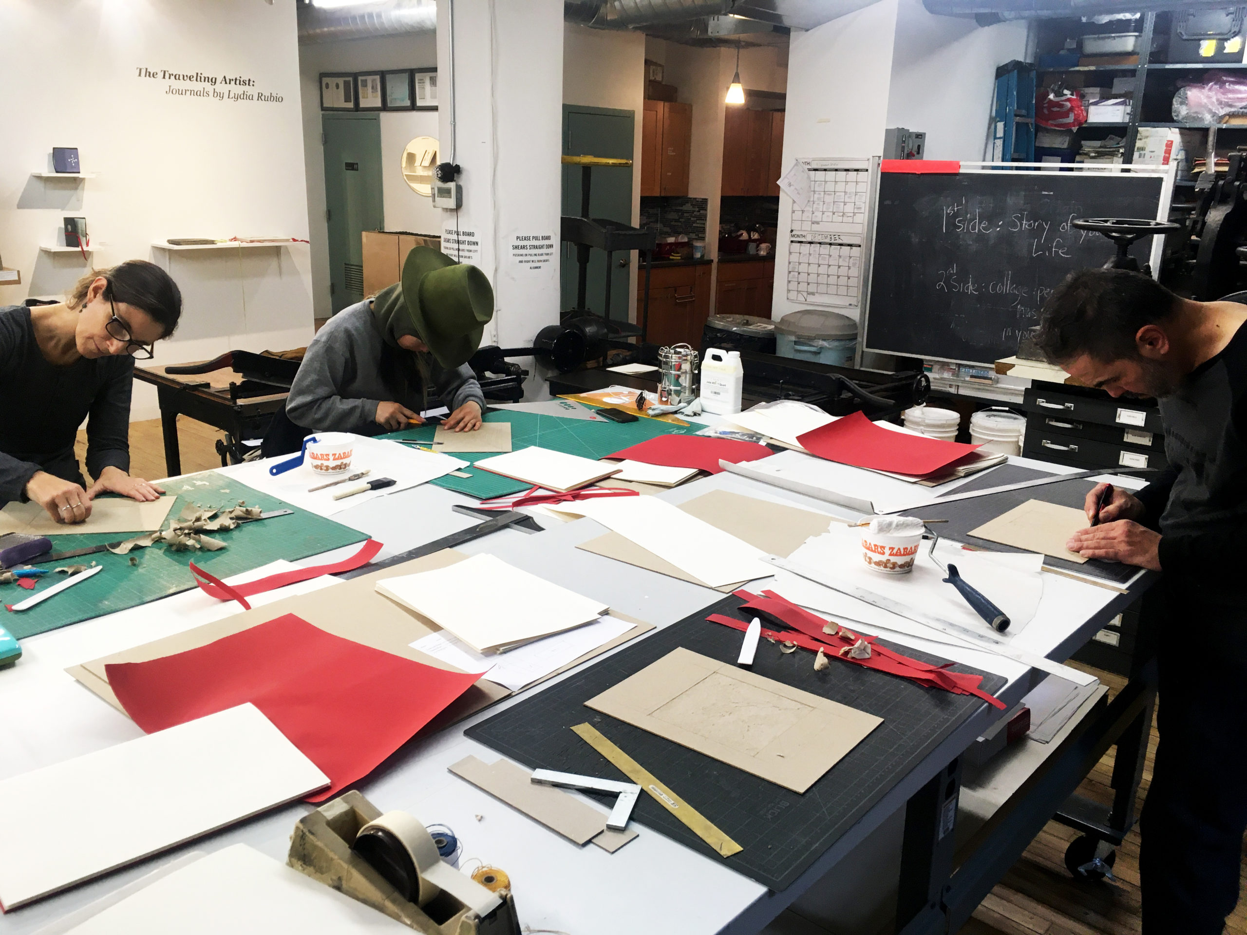 Three artists are cutting bookboard for an artists book. The table they're working at has supplies (paper, rulers, tape, etc) spread out all over it.