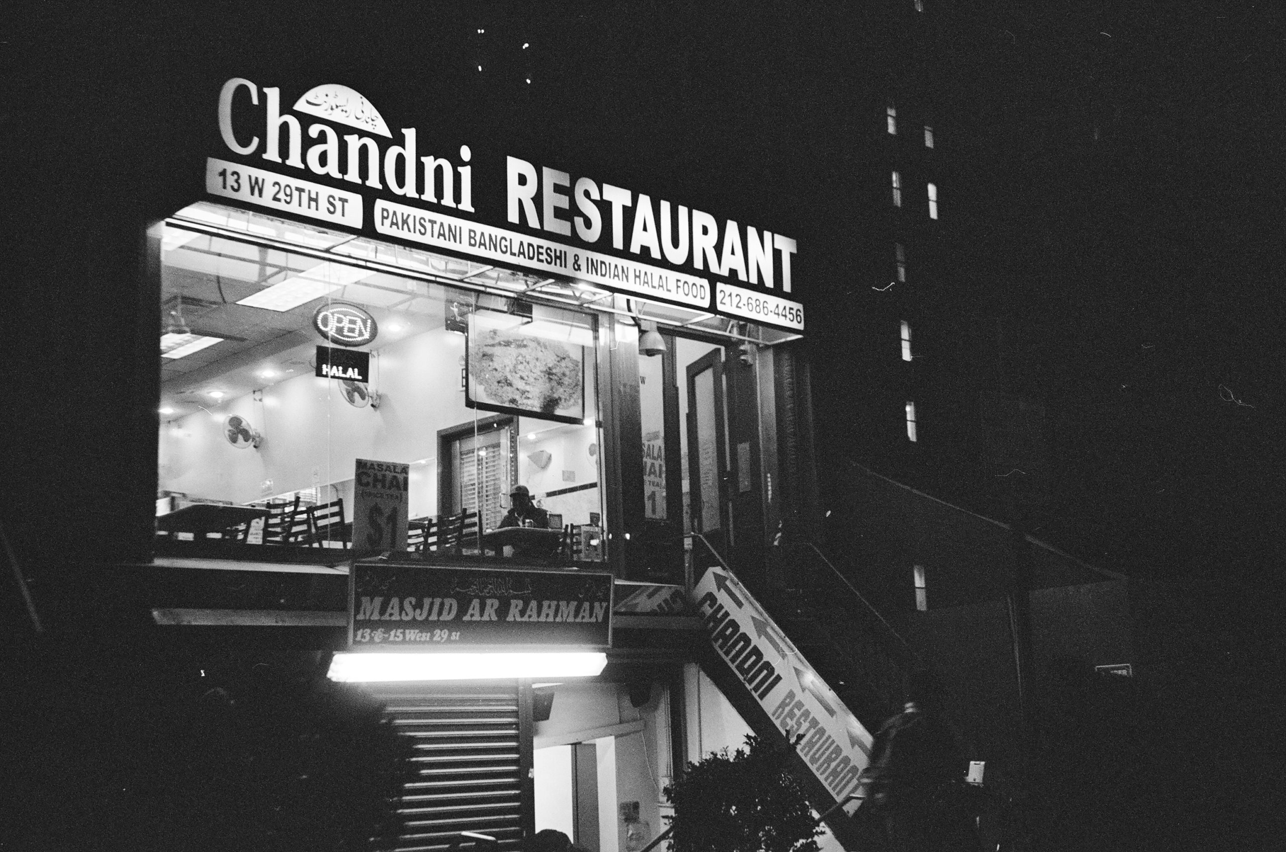 Black and white photo of Chandi Restaurant at night. There is a man in the window of the restaurant, he is seated at a table with his meal.