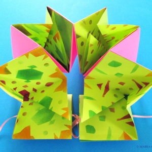 A bright green, orange, and pink flexagon book by Maria Pisano. The book is open in front of a blue background.