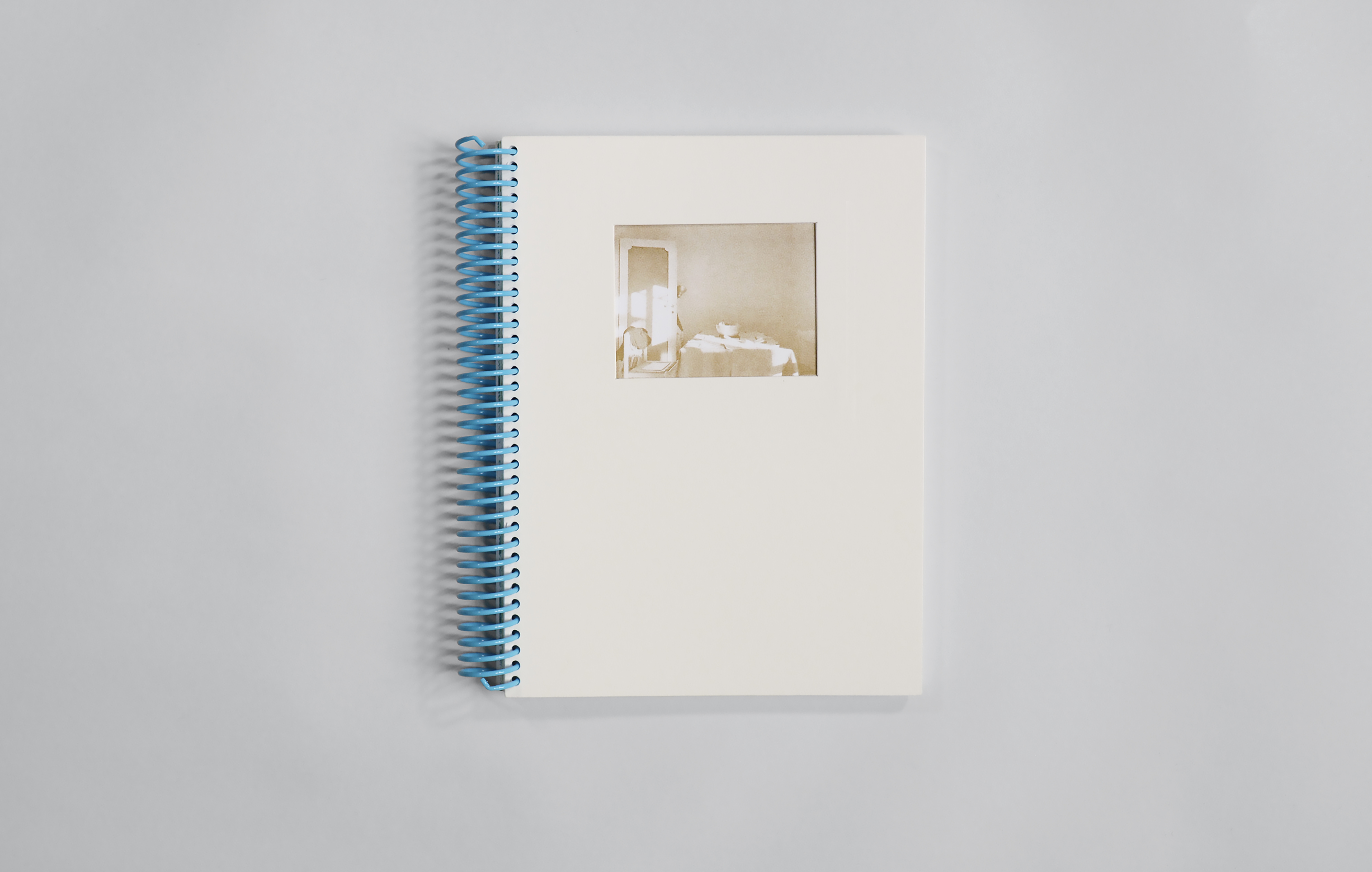 Front cover of exhibition catalog with die-cut window on white cardstock revealing brown risograph-printed image of a table and mirror. Bound with a blue spiral coil.