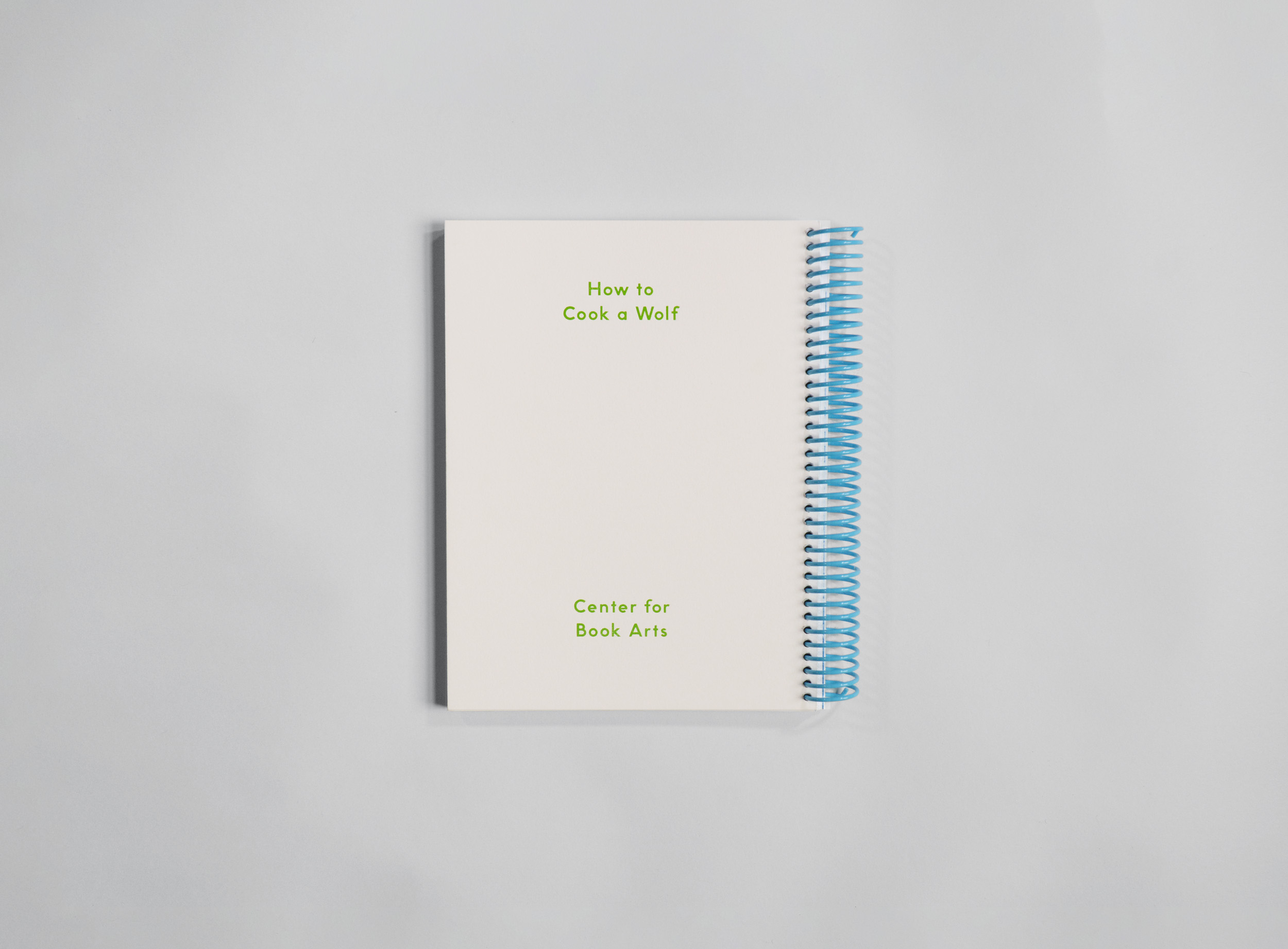 Back cover of exhibition catalog with green foil stamping that reads "How to Cook A Wolf / Center for Book Arts" on white paper, bound with a blue spiral coil.