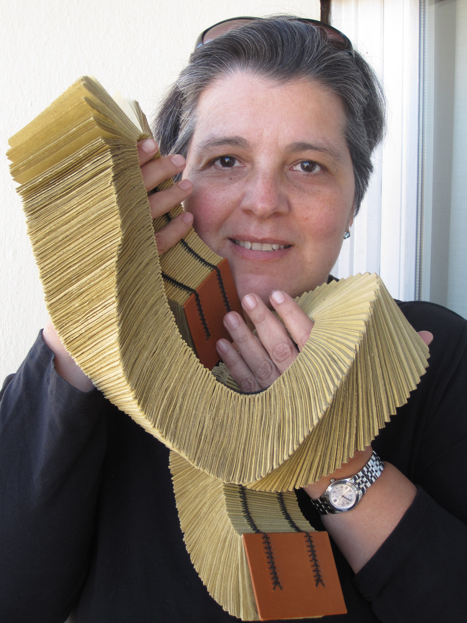 image of a woman with short hair holding an elaborately bound book, warped around her hands and against her face. She is staring into the camera with a smile