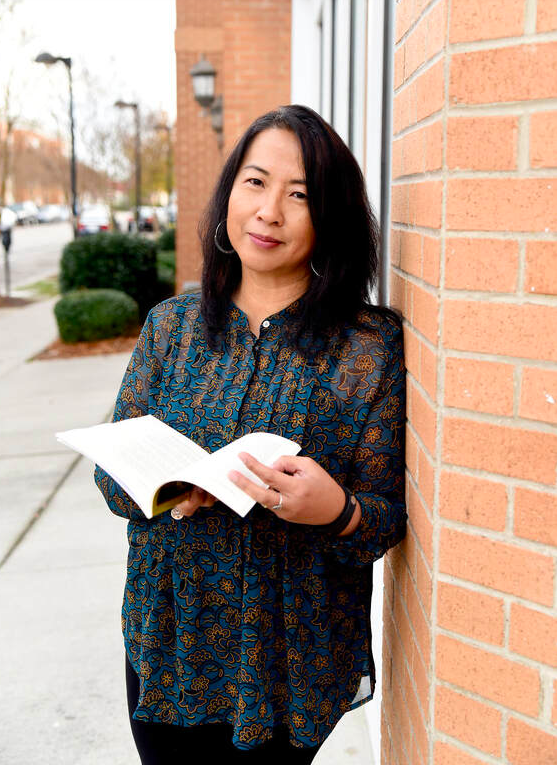 woman with dark hair leans against a brick wall holding an open book