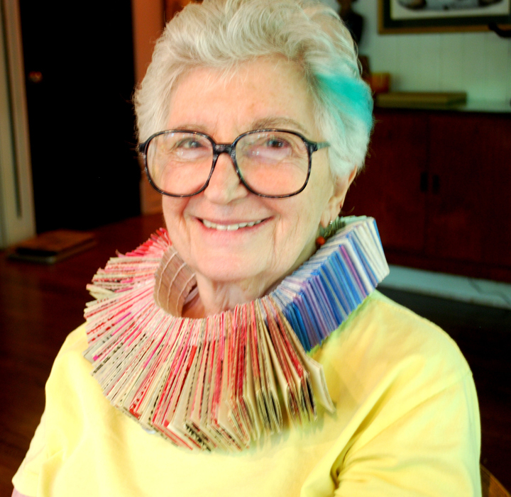 A photo of an elderly woman with cropped white hair and a bold paper necklace. She is smiling and wearing a yellow shirt.