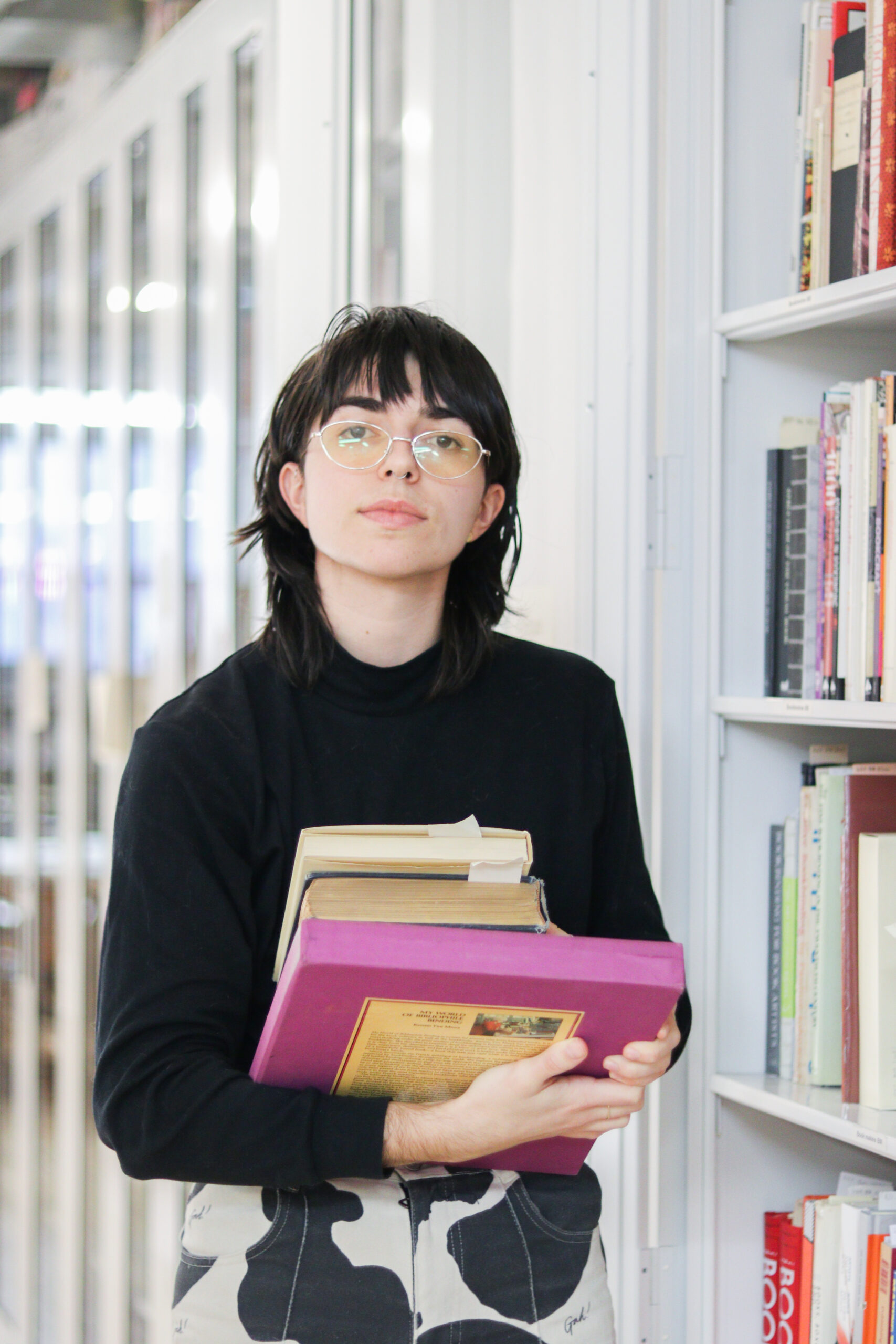 Gillian, a white person with shoulder-length brown hair and bangs, wearing a black turtleneck and cow print pants, stands in front of the CBA bookcases holding a stack of books and looking at the camera with a neutral expression.