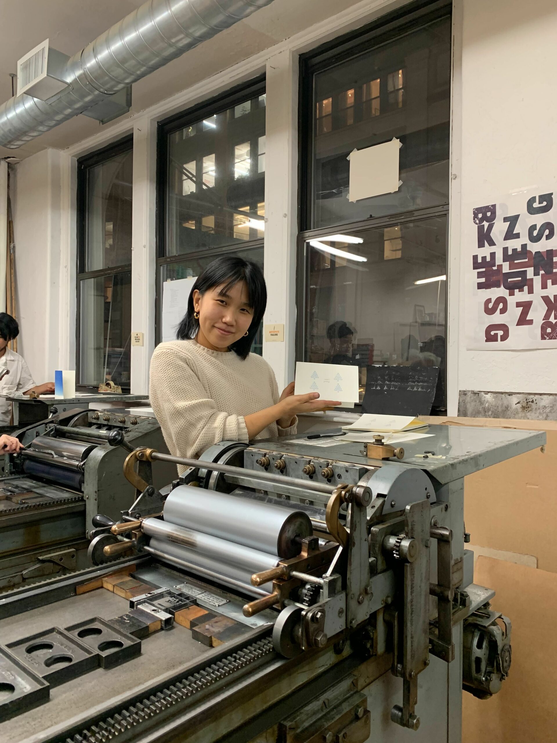 Caslon stands at a Vandercook printing press holding a printed card and smiling.