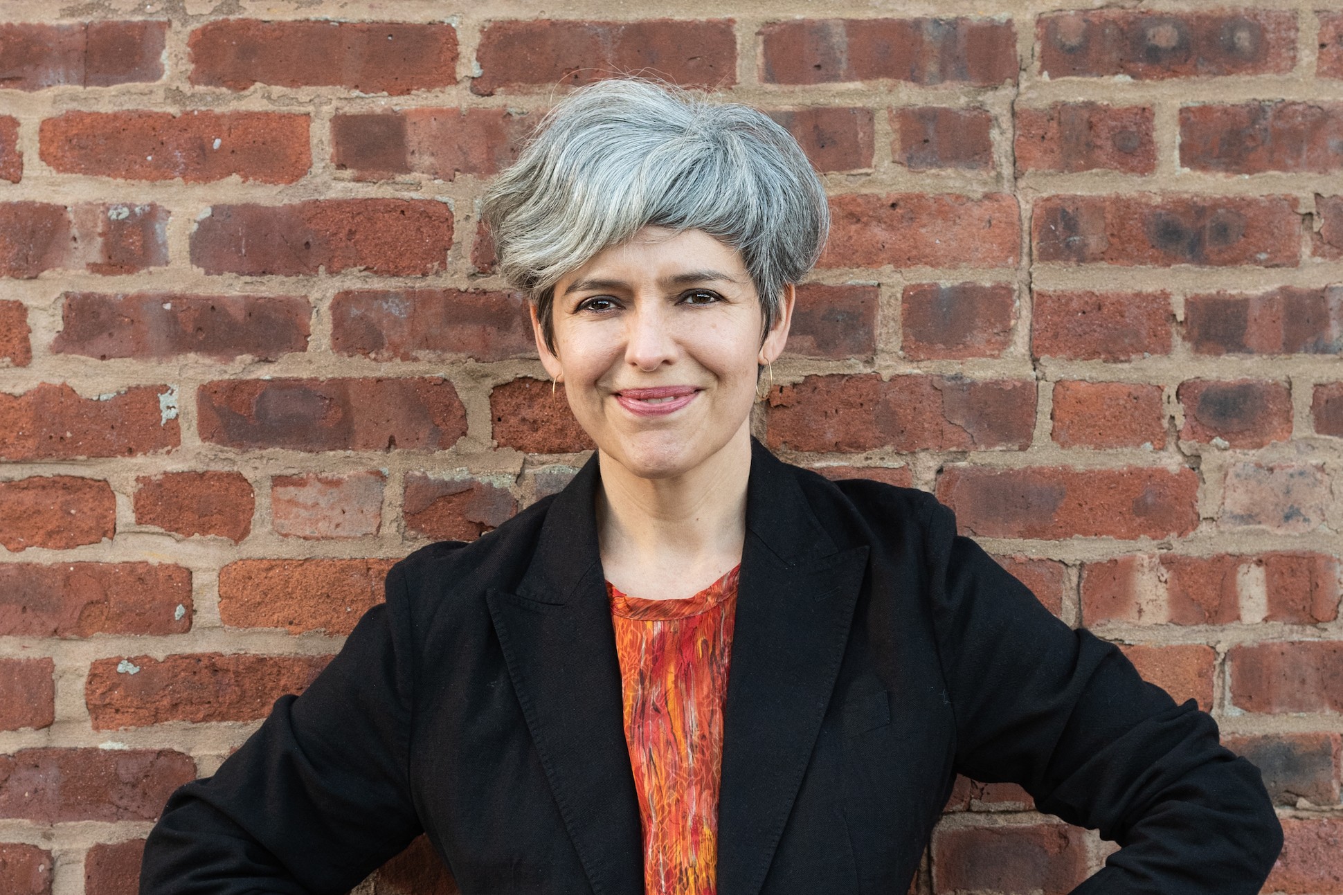 woman with short grey hair stands in front of a brick wall