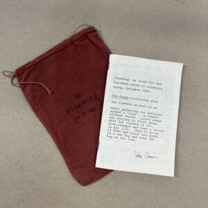 A white pamphlet with black text laying on top of a dark red or crimson pouch that has the words "a ripening" written on it. the pamhplet and pouch are photographed from above and are on a sandy gray background.