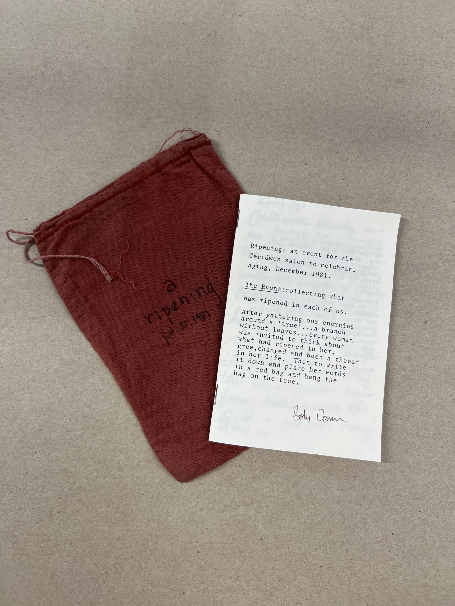 A white pamphlet with black text laying on top of a dark red or crimson pouch that has the words "a ripening" written on it. the pamhplet and pouch are photographed from above and are on a sandy gray background.