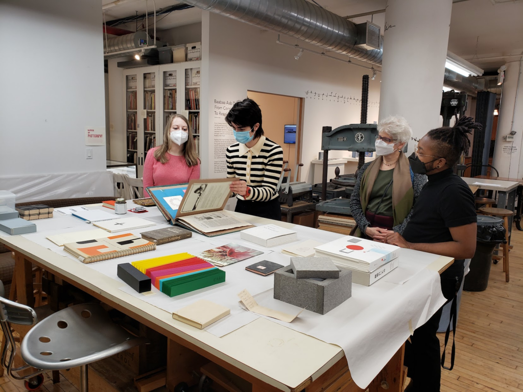 CBA's Librarian Gillian is showing a scrapbook to three visitors. They are wearing a striped sweater and a blue mask. The group is in the bindery of Center for Book Arts. In the foreground is a table full of books from the Fine Art Collection. 