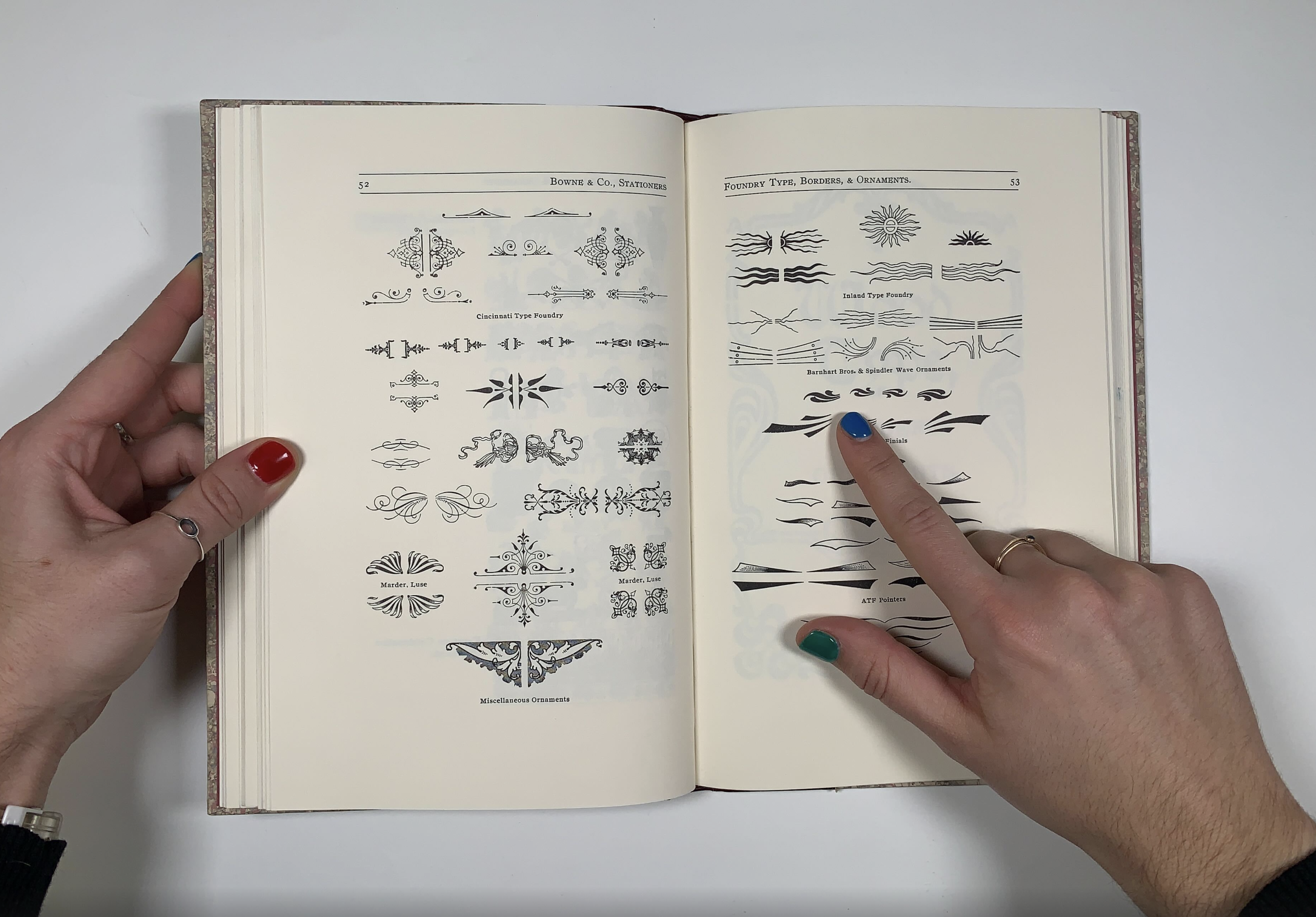 Two hands hold a type specimen book before a plain white background. The left hand holds the book open and the right hand points to a symbol on the page.