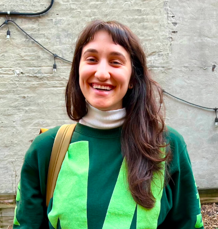 Pilar grins in front of a white-painted brick wall; she is wearing a green sweatshirt and has long brown hair.