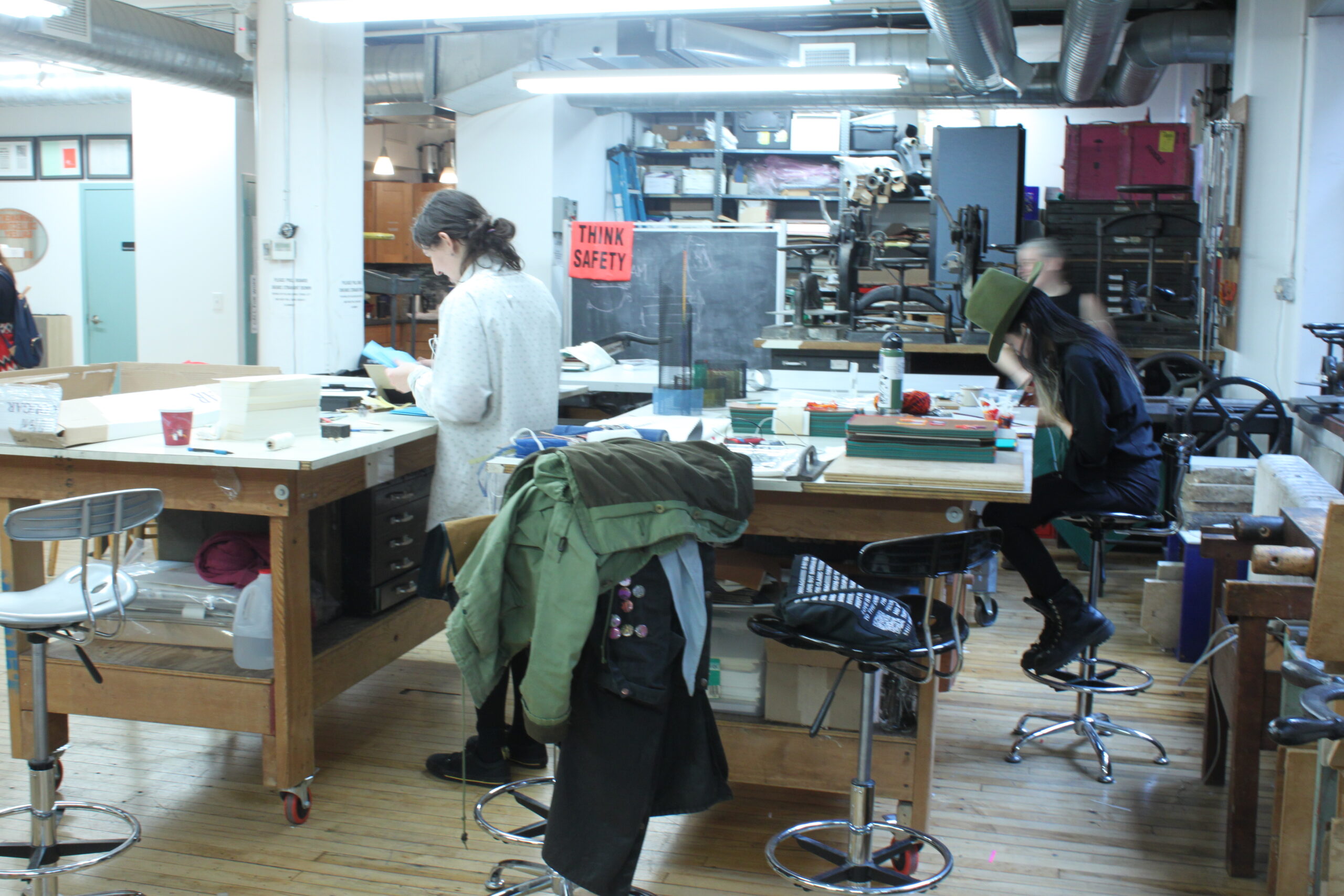 A view of the CBA bindery with two tables visible, Zoe is working at the left table and Linda is working at the table on the right.