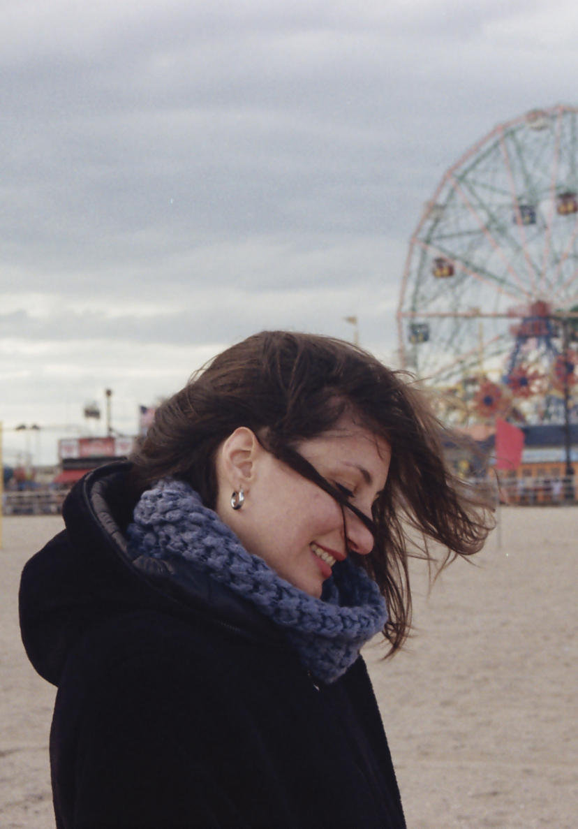 Caterina is in a jacket and scarf on a beach with her long brown hair blowing in the wind. She is smiling and there is a ferris wheel behind her.