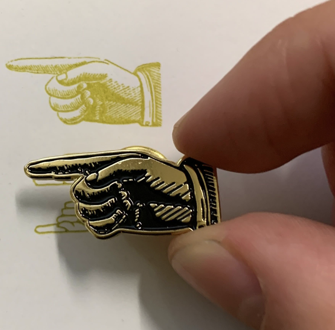 A gold pin shaped like a pointing hand, held in someone's fingers above a piece of paper printed with the same image of the pointing hand in chartreuse ink.