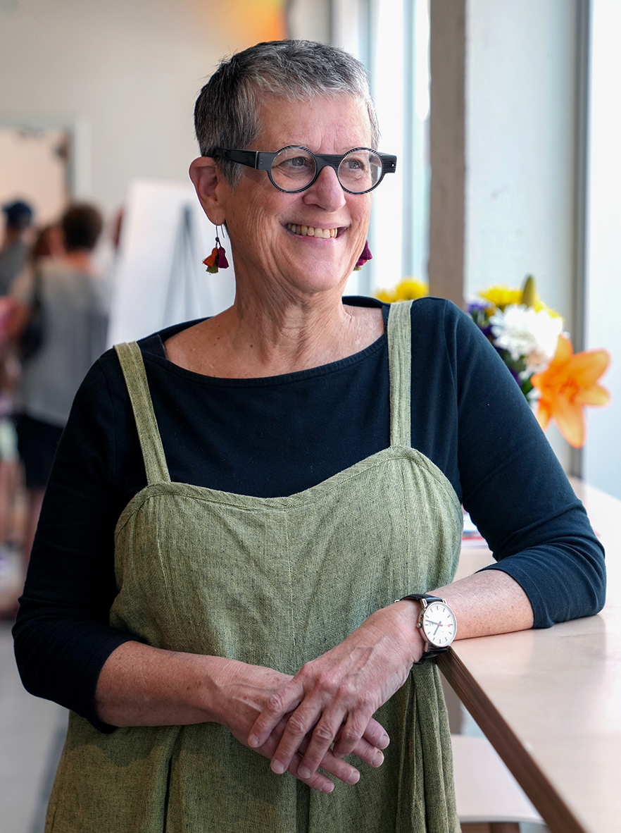 Lynne stands in a brightly lit studio space, leaning on a wooden table. She wears blue glasses, dangly earrings and green linen overalls over a dark blue long sleeve shirt. She has short hair and is smiling, looking beyond the camera.
