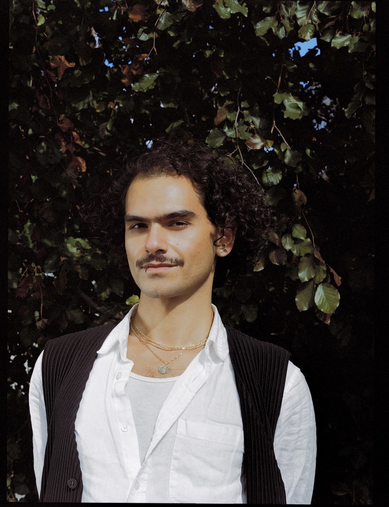 Darian standing before a leafy tree or shrub, wearing a white shirt with a dark vest